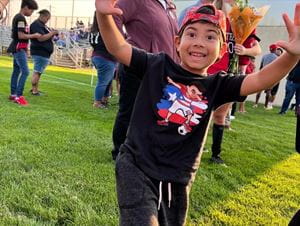 A child poses with arms outstretched and a big smile. He's wearing a backwards cap and is standing on a grass field.