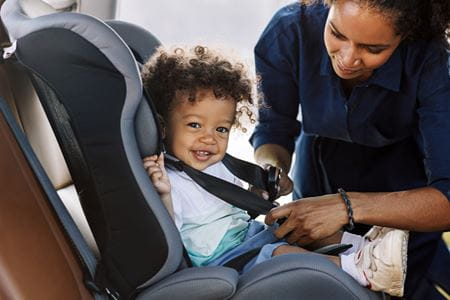 a child with dark curly hair smiles as they get buckled in to their car seat by a parent