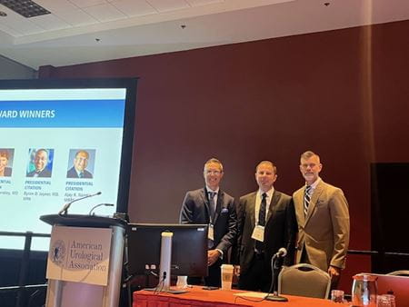 Nabil Adra stands behind a podium with colleagues Clint Cary and Tim Masterson at an American Urological Association event.