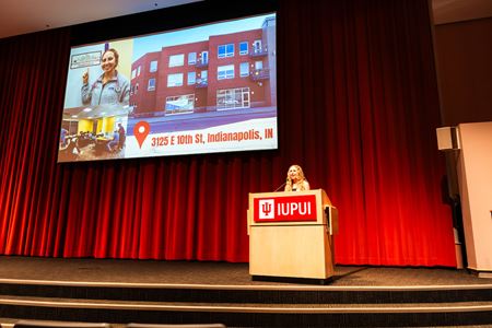 Elizabeth at an IUPUI podium on stage with a large screen behind showing the Student Outreach Clinic