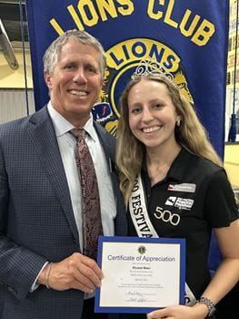 Elizabeth Baker with her dad in front of a Lions Club banner holding a certificate for speaking