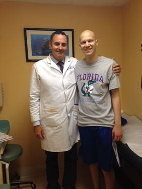 Miami pediatric oncologist Dr. Guillermo DeAngulo, in white coat, and Devin Gantzios-Cros, with no hair, stand side-by-side in a treatment room during his cancer treatment period in 2015.