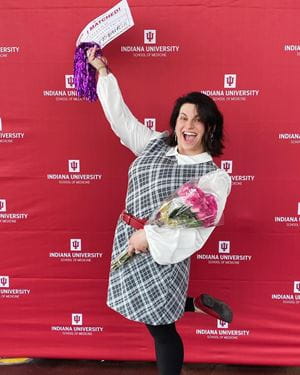 Hannah Kline wearing checked dress, holding a flower bouquet and Match Day sign, in front of IU School of Medicine logo backdrop