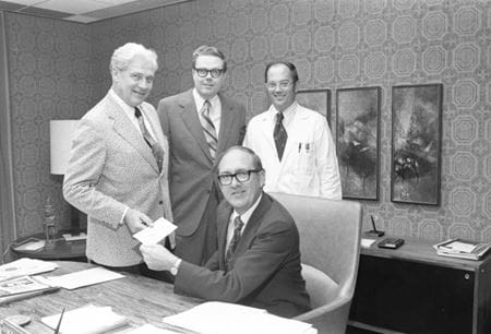 Chancellor Glenn Irwin, seated at a desk, receives a check from a donor as Walter Daly and a faculty physician observe