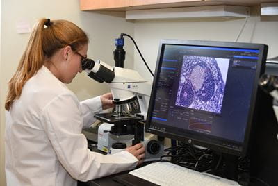 A female scientist looks into a microscope with a computer next to her showing the slide image