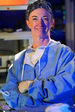 Portrait of Dr Jennifer Choi wearing surgical scrubs, mask around her neck, smiling with arms folded, in the surgery simulation laboratory