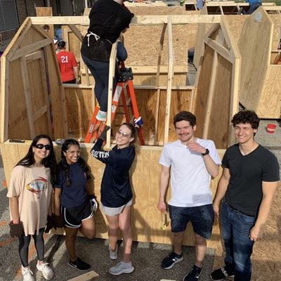 six students work to build sheds with habitat for humanity. one student is standing on a tall ladder while another holds the ladder safely and the other four students smile for the camera.