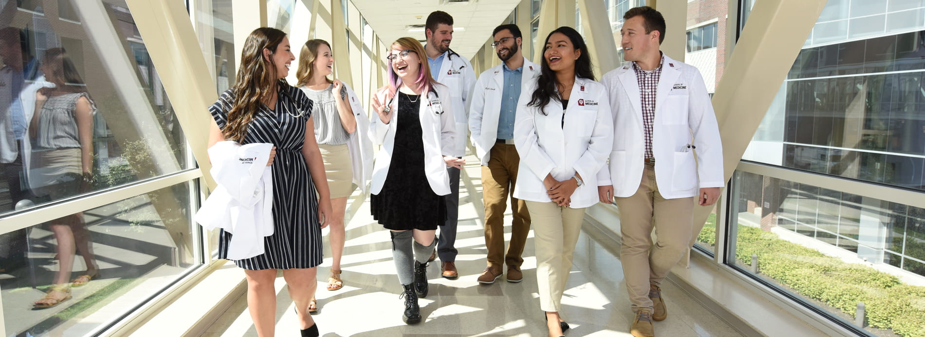 a diverse group of medical students in white coats smile and talk while walking down the hallway