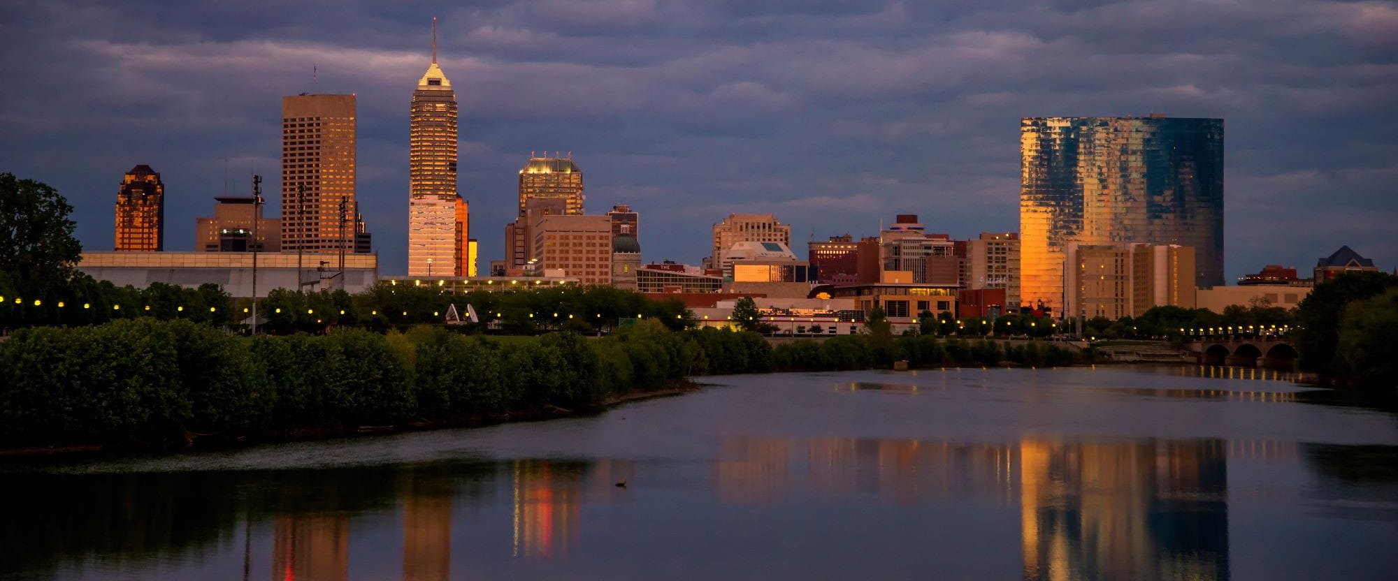 the indianapolis skyline is lit up at sunset. the white river flows gently in the foreground. There are moody dark clouds behind the buildings.