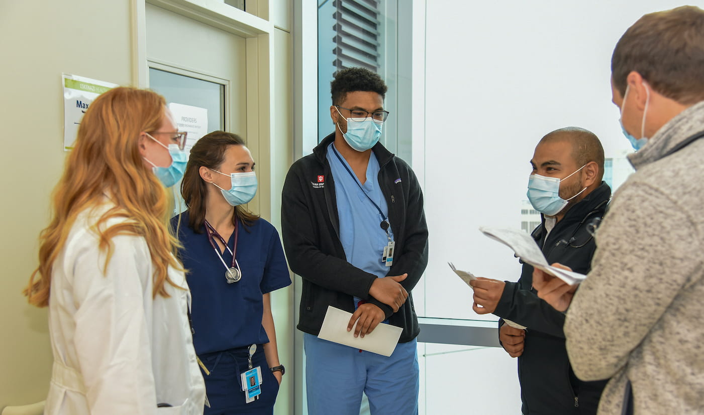 A mixed-gender group of young physicians in face masks consult each other in a brightly-lit hospital hallway.