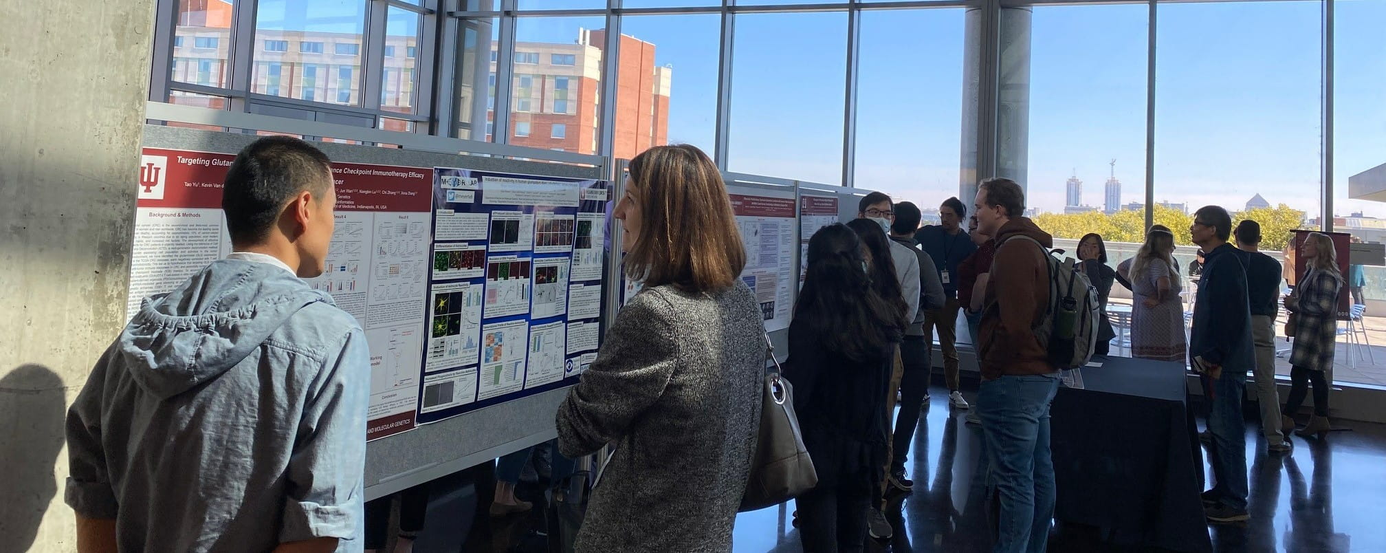 symposium attendees browse the poster session in front of a large window with the indy skyline in the background
