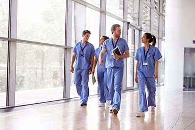 a group of physicians in blue scrubs walk down a hallway lined with floor to ceiling windows