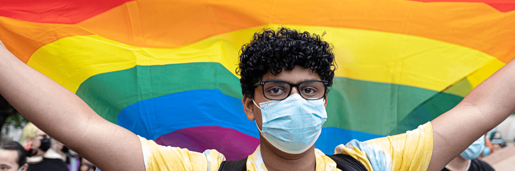 a young person with dark curly hair wearing a mask holds a large pride flag behind them
