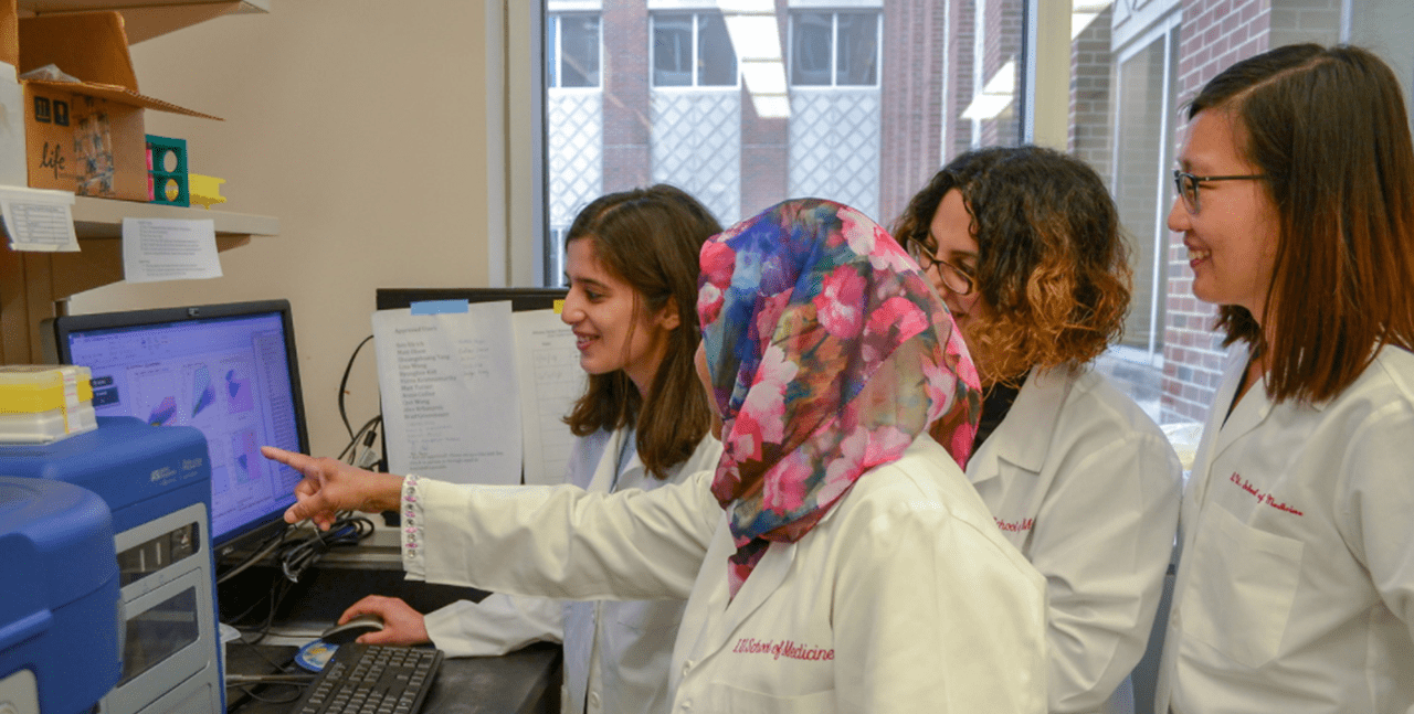a group of students wearing white coats review data on a computer in the lab