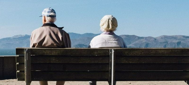two elderly people sit together on a bench and look at a beautiful view of the mountains