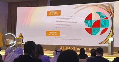 Dr. Barbara Maxwell stands infromt of a giant screen, presenting to an international audience of health care professionals and educators at All Together Better Health IX in Qatar