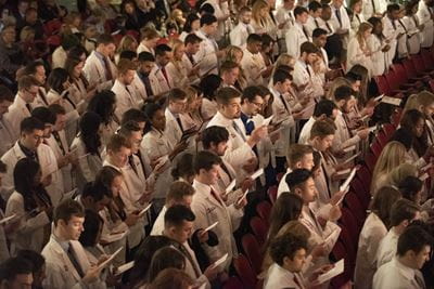 rows of medical students in white coats stand in an auditorium as they take the physician's oath