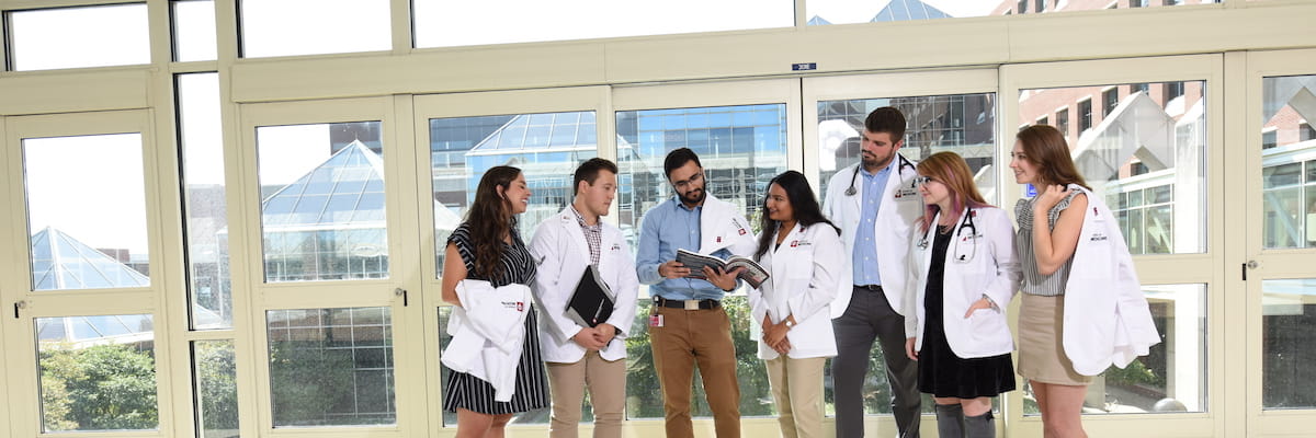 a diverse group of medical students stand in a semi circle holding and wearing white coats and looking at a book together